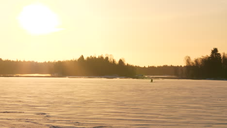 Wide-shot-of-distant-fisherman-on-frozen-lake,-ice-fishing-at-golden-sunrise-on-a-cold-winter-day