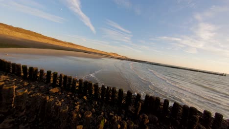 Fast-low-angle-view-above-a-dyke-and-a-beach-with-wave-breakers-during-low-tide