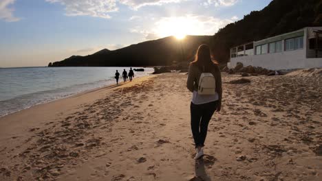 Woman-walks-along-beach-as-sun-sets-behind-the-mountains