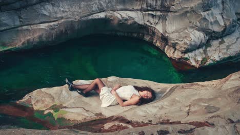4K-Cinemagraph---seamless-video-loop-of-a-young-brunette-model-woman-in-a-white-dress-lying-at-the-edge-of-a-gorge-by-a-blue-mountain-river