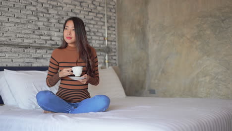 A-lovely-young-Asian-woman-sits-cross-legged-on-the-bed-sipping-a-cup-of-coffee-tea
