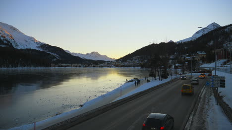 St-Moritz,-Switzerland-frozen-lake-and-road-with-light-traffic-and-people-walking-by