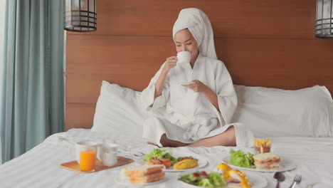 A-young-Asain-woman,-fresh-from-a-shower-dressed-in-a-robe-and-hair-wrapped-up-in-a-towel,-sips-from-a-coffee-cup-while-surveying-a-bounty-of-food-spread-out-on-her-hotel-room-bed