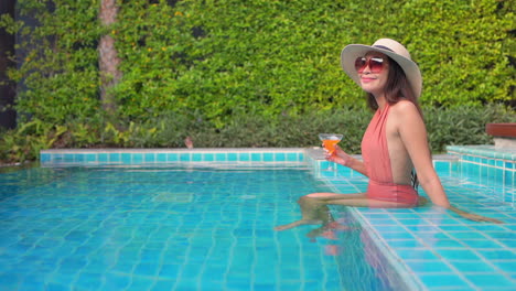 A-beautiful-woman-in-a-coral-bathing-suit,-sunglasses,-and-straw-sun-hat-sits-on-the-step-of-a-pool-while-drinking-a-colorful-tropical-drink