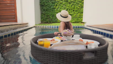 Asian-woman-in-mustard-color-bathing-suit-pulls-on-her-floating-tray-of-breakfast-inside-a-pool