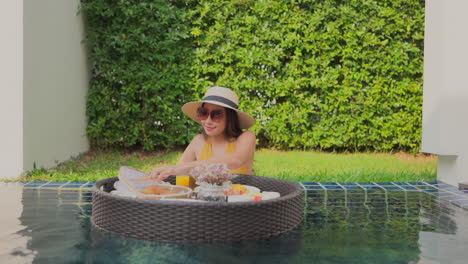 Attractive-Asian-woman-in-mustard-color-bathing-suit-enjoys-floating-tray-of-breakfast-inside-a-pool