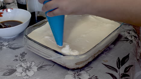 Baker-Applying-Fresh-Cream-Using-Piping-Bag-To-Tiramisu-Desert-Cake-On-Kitchen-Table