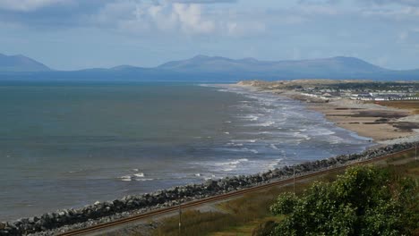 Looking-across-a-railway-line-through-to-Coastal-waves-and-beach-and-in-the-background-can-be-seen-the-mountains-of-the-Llyn-Peninsula,-Wales,-UK---10-Second-version