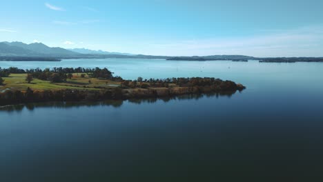 Chiemsee-lake-in-Bavaria,-Germany-with-the-river-delta-Tiroler-Ache-in-autumn-with-blue-water-and-sky,-trees-on-an-island-and-horizon-with-the-German-and-Austrian-alps-in-the-background