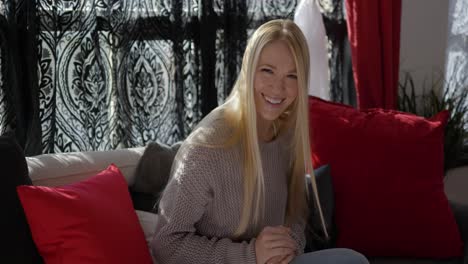 Blonde-white-caucasian-beautiful-girl-or-women-sitting-on-couch-with-red-pillows-in-front-of-window-curtains-with-natural-day-light-shining-through-as-she-smiles-waves-and-interacts-with-camera