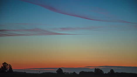 Wispy-clouds-cross-the-sky-above-the-wooded-horizon-at-dawn---wide-angle-sunrise-time-lapse