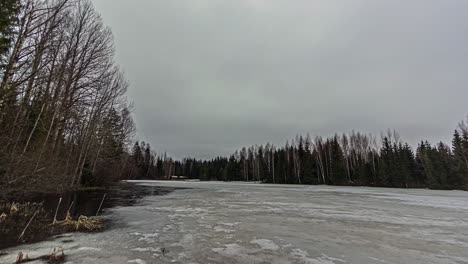 Melting-ice-of-frozen-forest-lake-and-moody-grey-sky,-fusion-time-lapse