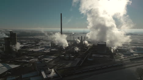 Smoke-and-steam-rising-of-an-industrial-facility,-CO2-emissions,-air-pollution,-exhaust-gases,-orbiting-aerial-drone-shot,-backlit-scene