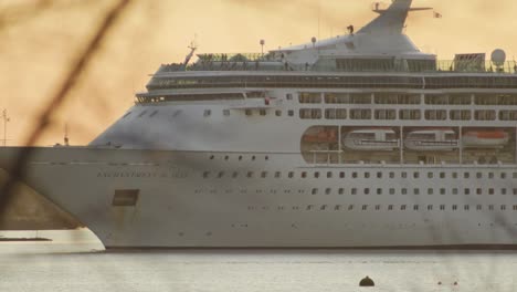 Cruise-ship-exits-port-at-sunset,-passengers-moving-on-deck-medium-telephoto-shot
