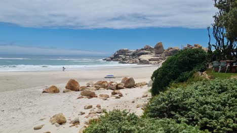 A-surfer-walking-on-a-perfect-looking-white-sand-beach-with-blue-water-and-some-rock-formations-in-the-background-near-Cape-Town