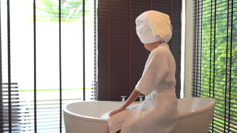 Young-Lady-in-a-white-bath-gown-sitting-on-a-bath-tube-side-and-taking-foam-with-her-hand-in-luxury-hotel-in-Indonesia