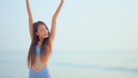 Beautiful-Asian-woman-model-standing-in-front-of-the-sea-looking-aside-interlock-her-fingers-and-raise-hands-up-and-spread-arms-over-her-head