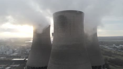 Aerial-slow-dolly-right-across-power-station-cooling-towers-smoke-steam-emissions-as-sunrise-hides-behind-tower