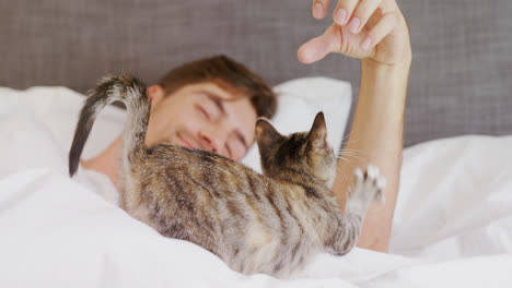 Glücklicher-Junger-Mann,-Der-Auf-Dem-Bett-Liegt-Und-Mit-Seiner-Getigerten-Katze-Spielt-4k-4k