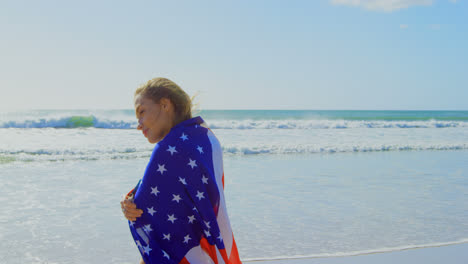 Seitenansicht-Einer-Jungen-Kaukasischen-Frau,-Die-In-Eine-Amerikanische-Flagge-Gehüllt-Ist,-Am-Strand-4k