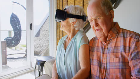 Front-view-of-Caucasian-senior-woman-using-virtual-reality-headset-and-senior-man-siting-on-sofa-at-