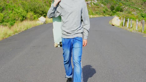 Side-view-of-cool-young-caucasian-man-with-skateboard-walking-at-countryside-road-4k