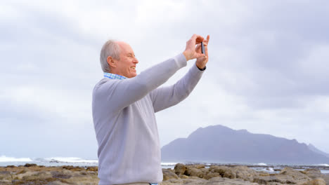 Front-view-of-old-caucasian-senior-man-clicking-photo-with-mobile-phone-at-beach-4k