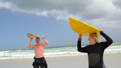 Front-view-of-old-caucasian-senior-couple-carrying-surfboard-on-her-head-at-beach-4k
