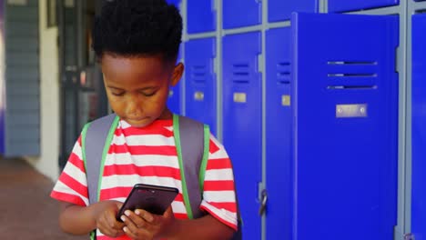 Front-view-of-African-American-schoolboy-with-schoolbag-using-mobile-phone-in-school-corridor-4k