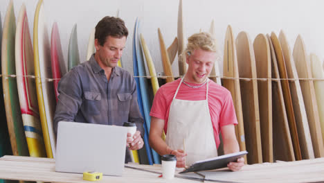 Two-Caucasian-male-surfboard-makers-working-on-projects-using-a-laptop-computer