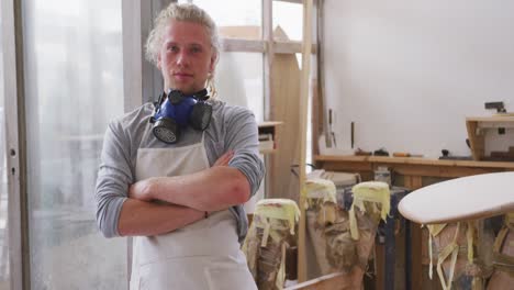 Caucasian-male-surfboard-maker-wearing-a-face-mask-and-standing-in-his-studio-with-his-arms-crossed