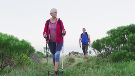 Senior-hiker-couple-with-backpacks-and-hiking-poles-while-walking-in-the-grass-field.