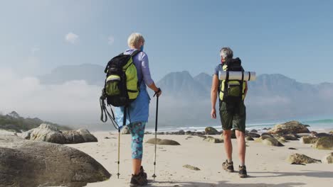 Rear-view-of-senior-hiker-couple-wearing-face-mask-with-backpacks-and-hiking-poles