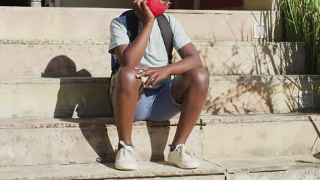 Sad-african-american-boy-wearing-face-mask-sitting-on-stairs-outdoors-on-a-sunny-day