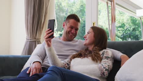 Kaukasisches-Paar-Lächelt,-Während-Es-Einen-Videoanruf-Auf-Dem-Smartphone-Führt-Und-Zu-Hause-Auf-Der-Couch-Sitzt