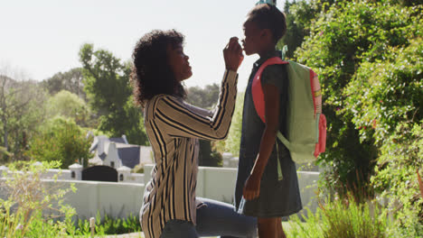 African-american-mother-putting-on-face-mask-on-daughter-with-backpack-outdoors-on-a-bright-sunny-da