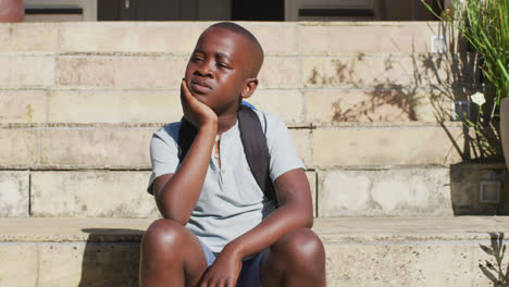 Trauriger-Afroamerikanischer-Junge-Mit-Gesichtsmaske,-Der-An-Einem-Sonnigen-Tag-Auf-Der-Treppe-Im-Freien-Sitzt
