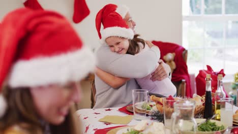 Kaukasisches-Mädchen-Mit-Weihnachtsmütze-Umarmt-Ihren-Vater,-Während-Es-Am-Esstisch-Sitzt-Und-Gemeinsam-Das-Mittagessen-Genießt