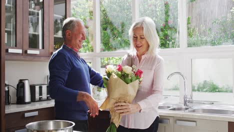 Älterer-Mann-Schenkt-Seiner-Frau-Einen-Blumenstrauß-Und-Umarmt-Sie-Zu-Hause-In-Der-Küche