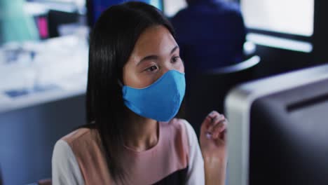 Asian-woman-wearing-face-mask-using-computer-while-sitting-on-her-desk-at-modern-office