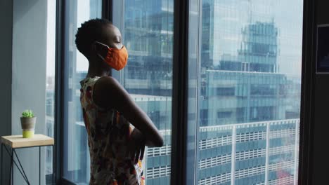 Thoughtful-african-american-woman-wearing-face-mask-looking-out-of-window-at-modern-office