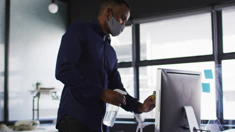 African-american-man-wearing-face-mask-cleaning-his-computer-with-disinfectant-at-modern-office