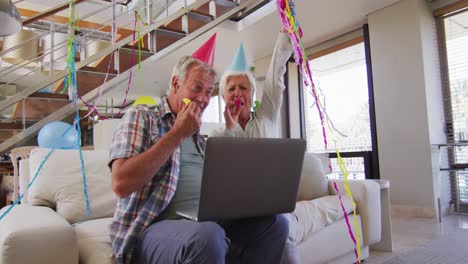 Senior-caucasian-couple-blowing-party-blowers-celebrating-birthday-while-having-a-video-call-on-lapt