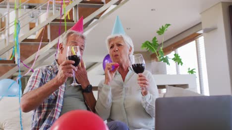 Senior-caucasian-couple-blowing-party-blowers-celebrating-birthday-toasting-wine-glasses-at-home