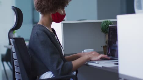 Mixed-race-woman-wearing-face-mask-in-office,-using-laptop