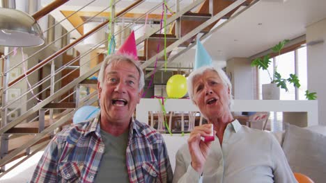 Senior-caucasian-couple-blowing-party-blowers-celebrating-birthday-enjoying-at-home