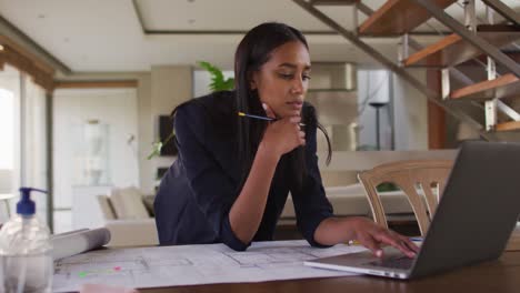 Mixed-race-woman-by-desk-at-home-using-laptop-looking-through-blueprints