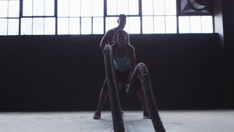 African-american-woman-exercising-battling-ropes-in-empty-urban-building-with-man-cheering-her-on