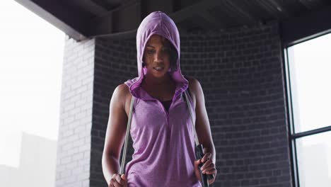 Portrait-of-african-american-woman-holding-skipping-rope-wearing-hoodie-looking-at-camera-in-an-empt