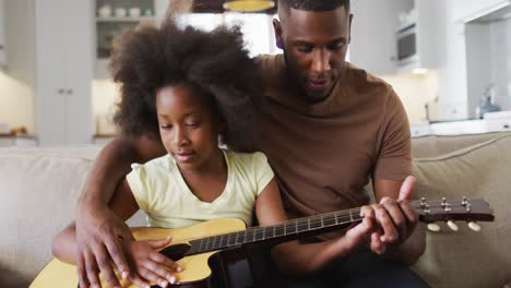 African-american-father-and-his-daughter-sitting-on-couch-playing-guitar-together